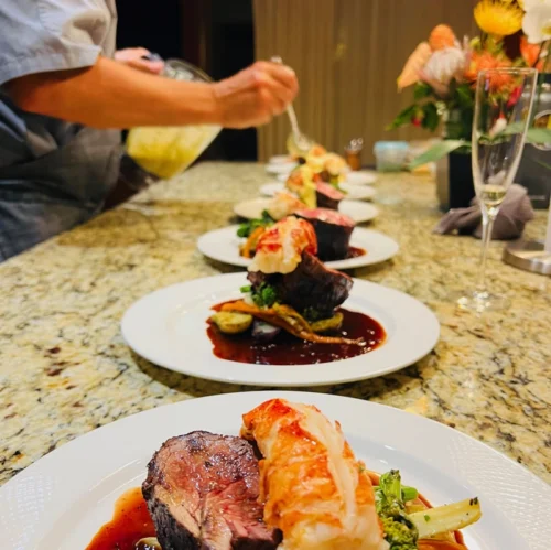 Plated steak and lobster dinners being finished with hollandaise by a chef.